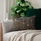 A rectangular lumbar cushion with an ornate design featuring birds, fruit, and leaves on a dark background, inspired by William Morris's art.