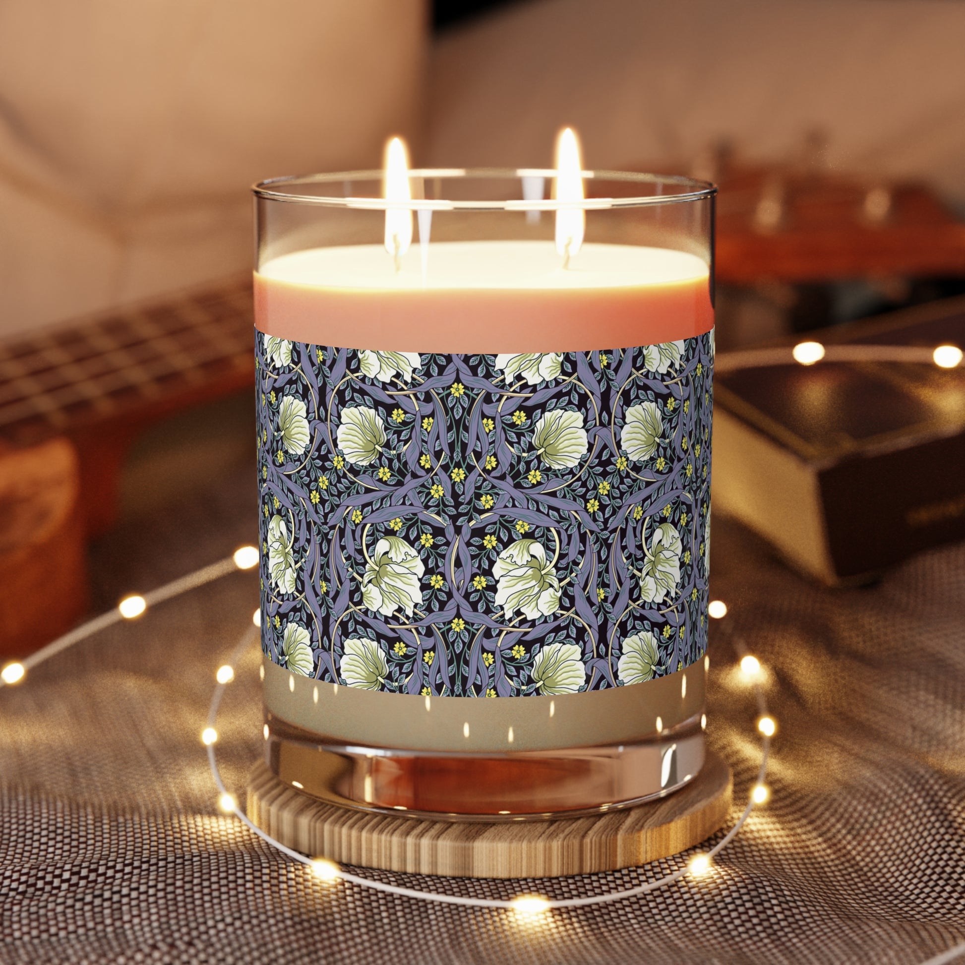 A clear candle with a lavender and sage William Morris & Co design called Pimpernel on the glass lid, alongside a brown cardboard lid with product information.