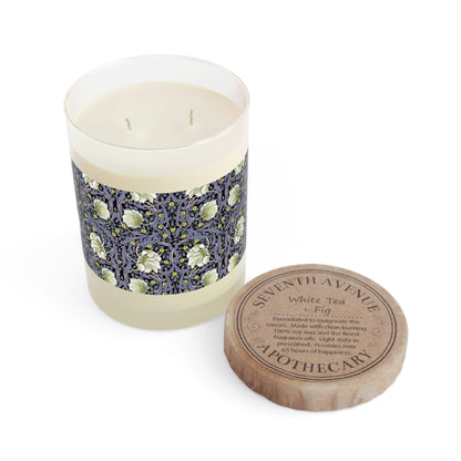 A clear candle with a lavender and sage William Morris & Co design called Pimpernel on the glass lid, alongside a brown cardboard lid with product information.