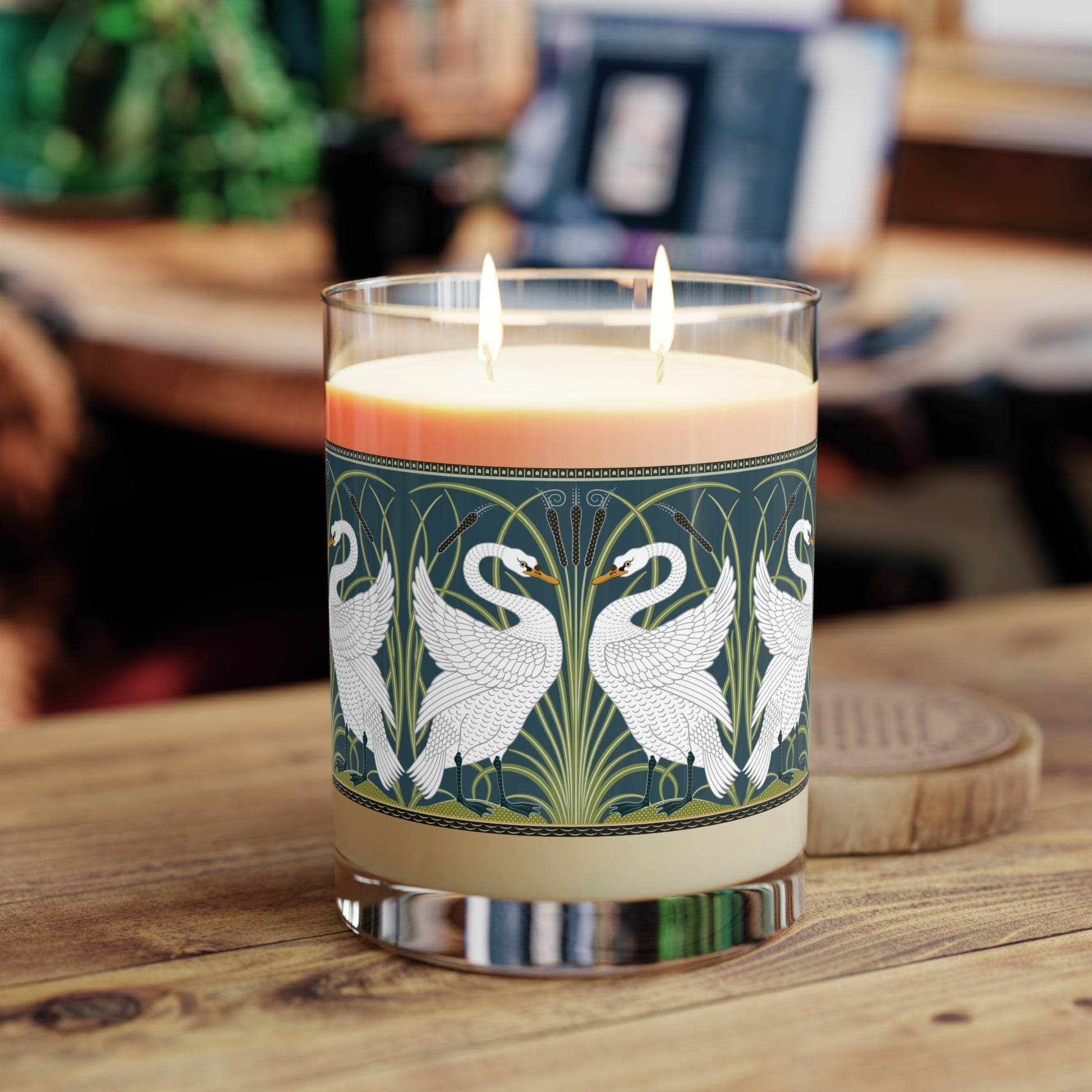 A clear glass candle with a white swan design on the label, alongside a brown cardboard lid with text.