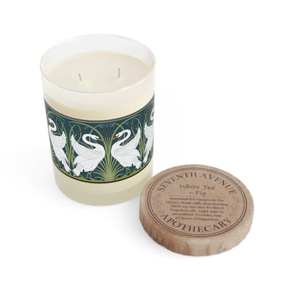 A clear glass candle with a white swan design on the label, alongside a brown cardboard lid with text.