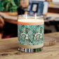 A two-wick candle in a clear glass jar sits on a wooden table. The candle has a cream-colored wax base and a decorative label featuring a teal and green botanical pattern with white flowers.