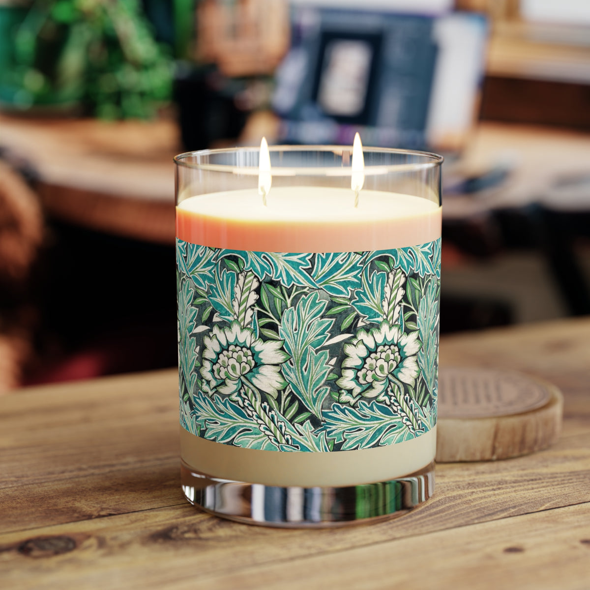 A two-wick candle in a clear glass jar sits on a wooden table. The candle has a cream-colored wax base and a decorative label featuring a teal and green botanical pattern with white flowers.