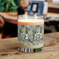 A double-wick scented candle in a clear glass jar sits on a wooden table. The candle has a cream-coloured wax body with a botanical patterned label featuring green and white leaves