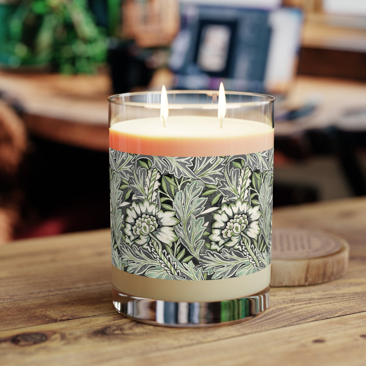 A double-wick scented candle in a clear glass jar sits on a wooden table. The candle has a cream-coloured wax body with a botanical patterned label featuring green and white leaves