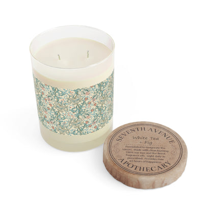 A beige candle with a floral pattern on the jar lid, labeled 'Seventh Ave White Tea & Fig', placed next to its cardboard lid.
