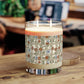 A lit two-wick candle in a clear glass jar sits on a wooden table. The candle has cream-colored wax and a decorative paper label featuring a beige grid pattern with blue birds and brown floral motifs.