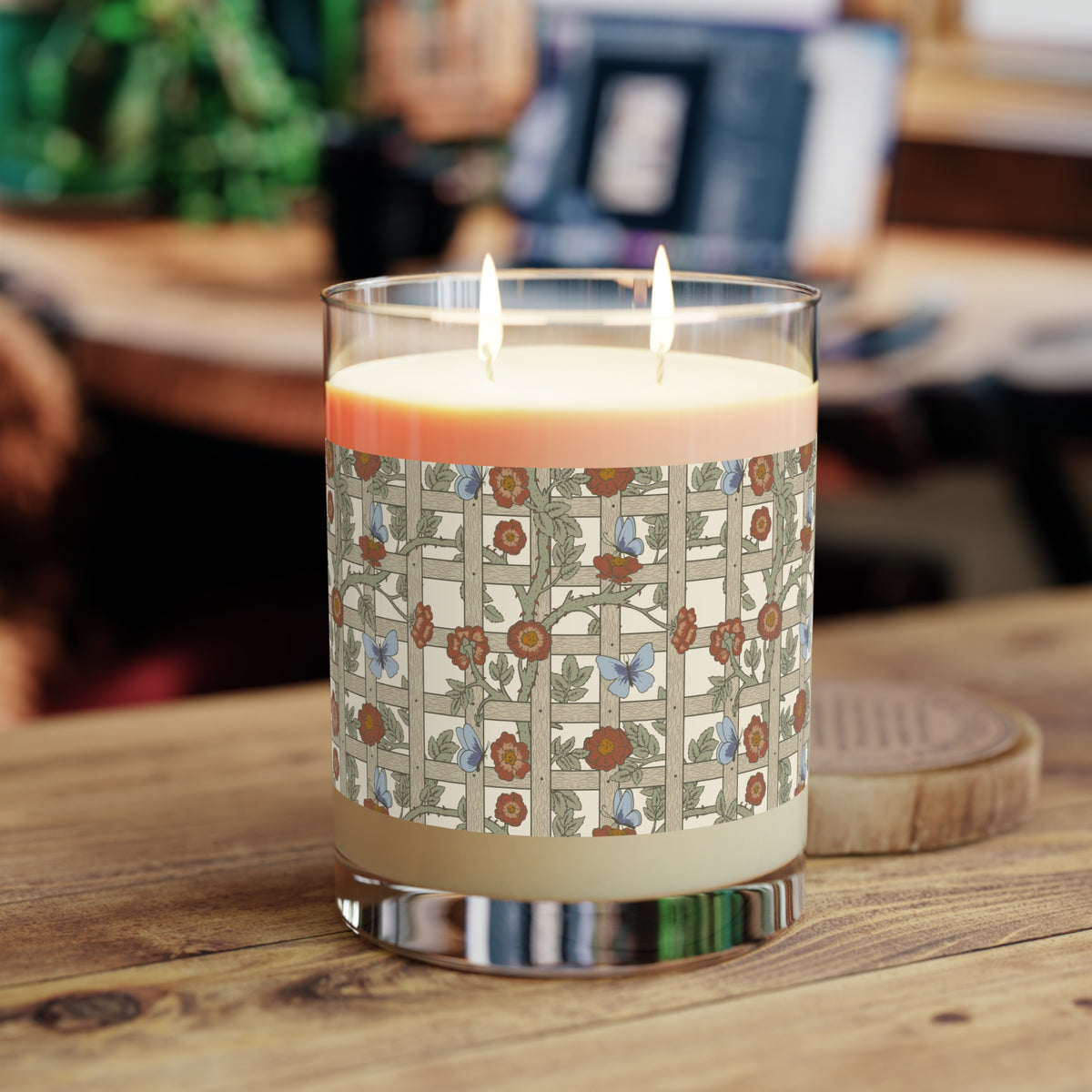 A lit two-wick candle in a clear glass jar sits on a wooden table. The candle has cream-colored wax and a decorative paper label featuring a beige grid pattern with blue birds and brown floral motifs.