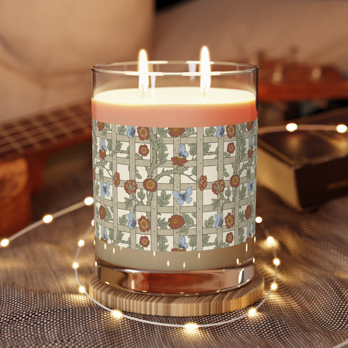 A three-wick candle in a clear glass jar with a warm pink wax and a floral patterned label, styled on a textured blanket with fairy lights and a guitar in the background.