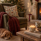 A cozy holiday living room scene with an olive green armchair, a red throw blanket, a patterned pillow, a lit candle on a wooden table, a sleeping dog on the rug, a Christmas tree, fireplace, stockings, and wrapped gifts.