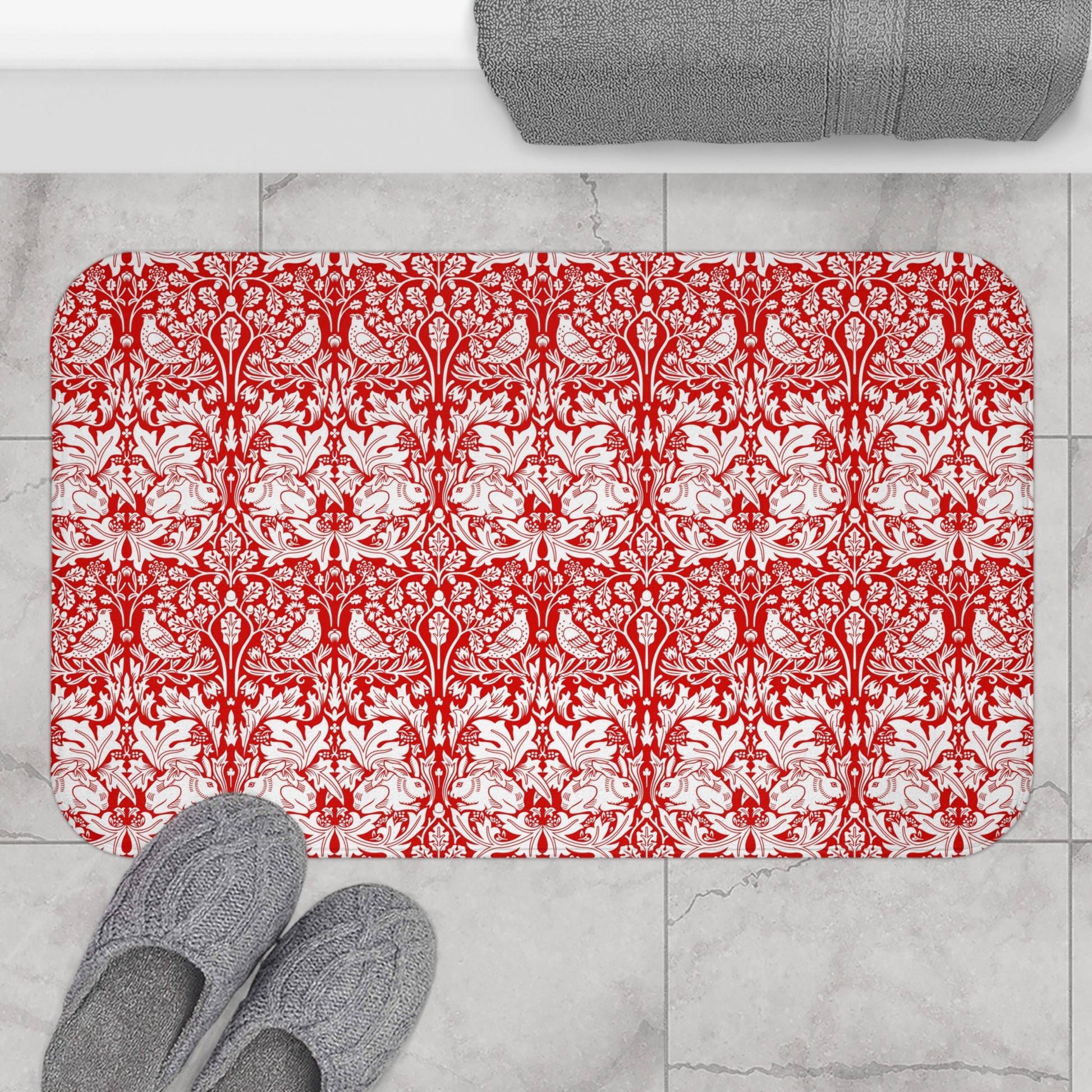 Red and white patterned bath mat on a tiled floor with gray slippers and towel.