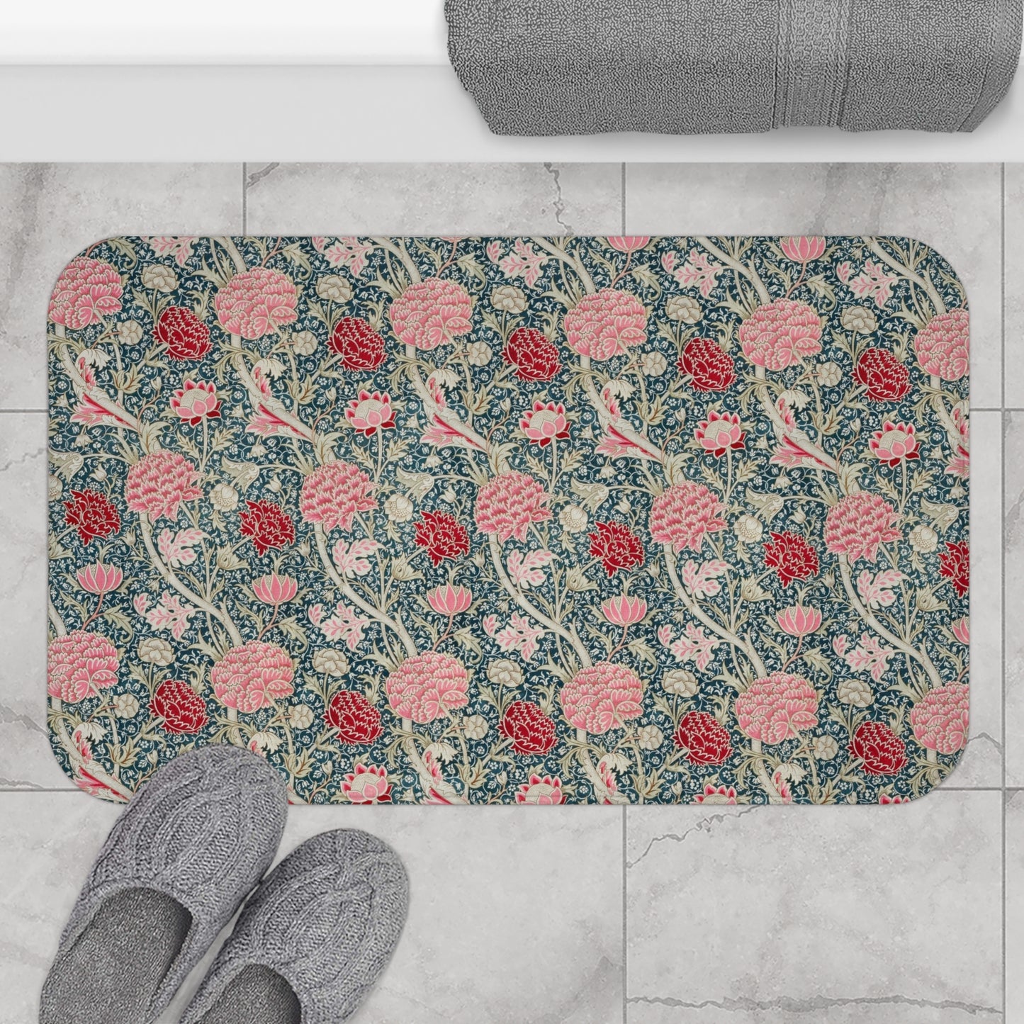 Floral patterned bath mat on a tiled floor with slippers and a towel.