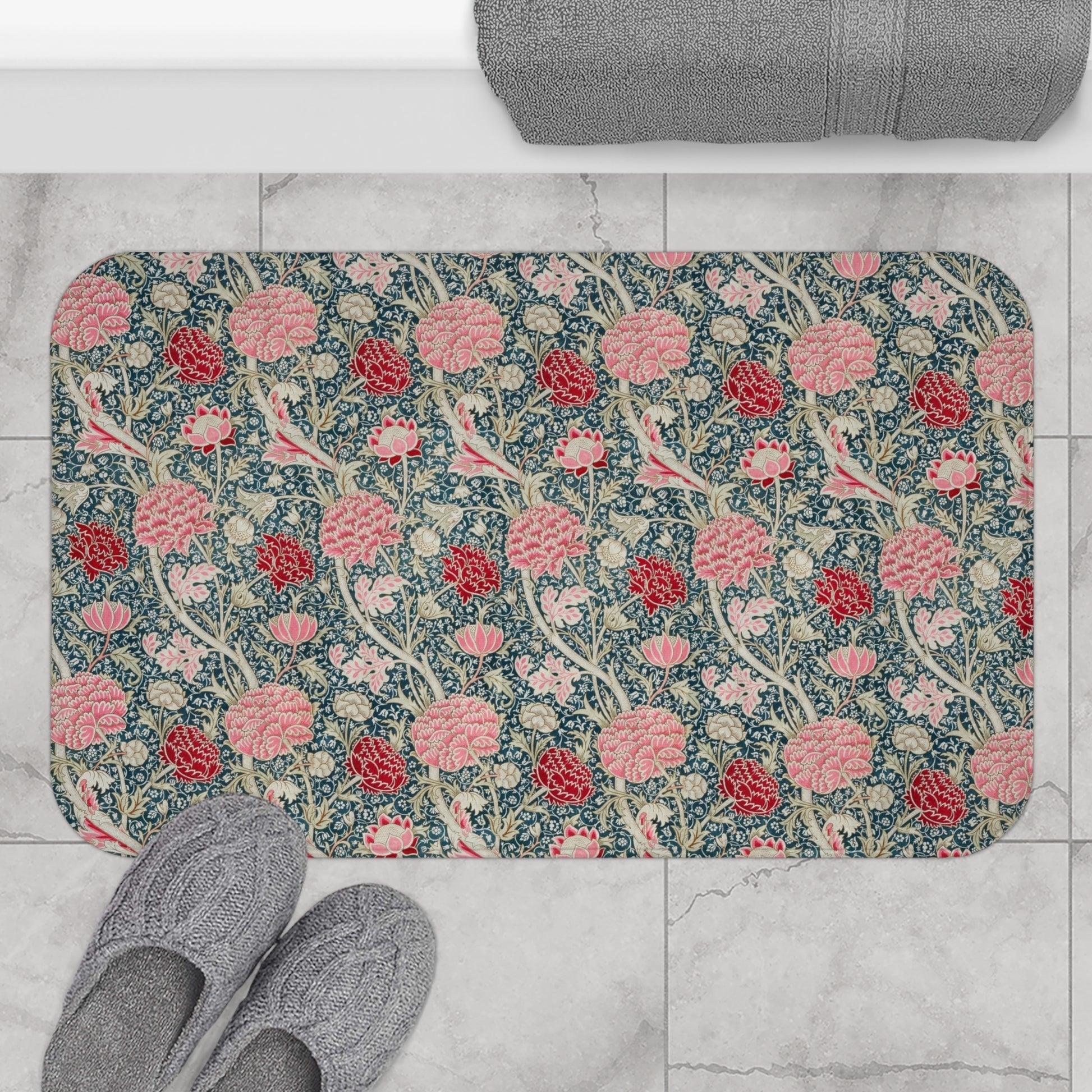 Floral patterned bath mat on a tiled floor with slippers and a towel.