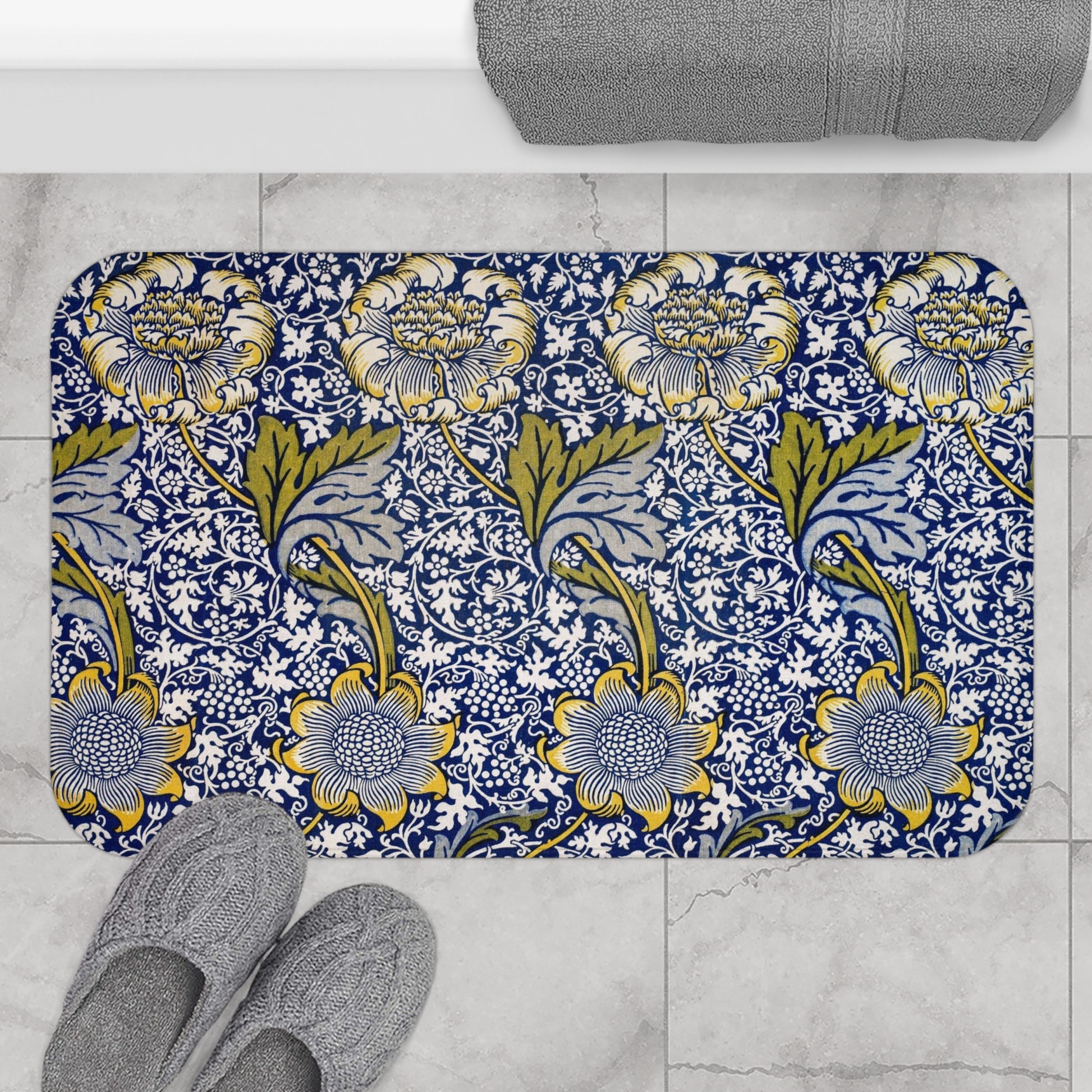 Floral patterned bath mat on a tiled floor with slippers and a towel.
