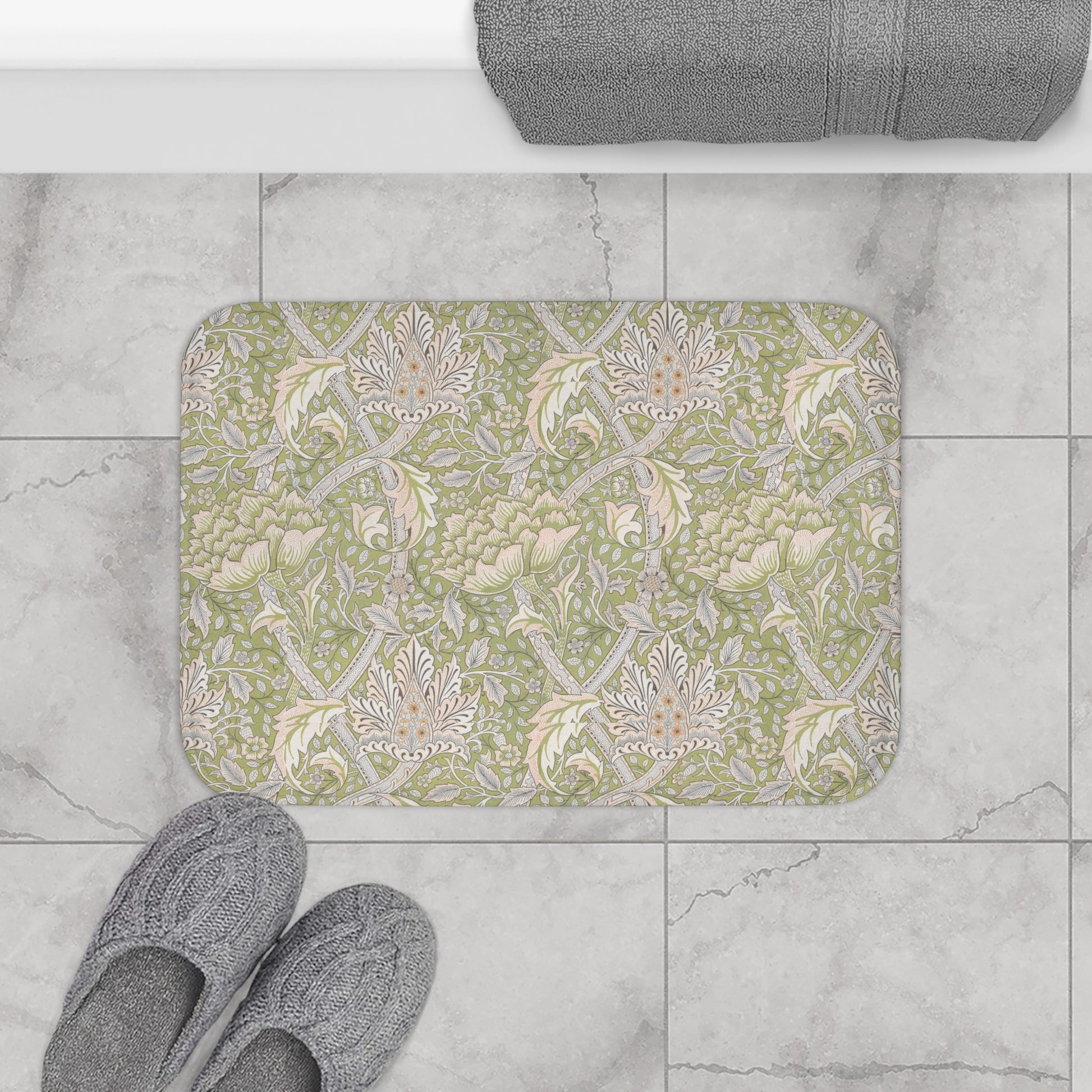 Decorative bath mat with floral pattern on a tiled floor, next to slippers and a towel.