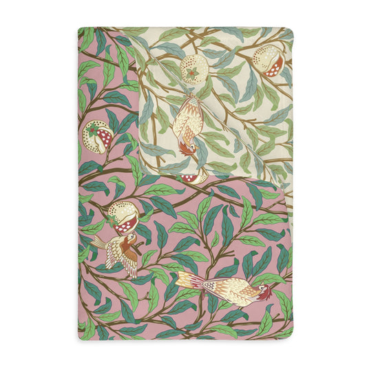 A rectangular blanket with a two-sided print featuring birds and pomegranate leaves on a pink background, inspired by William Morris.