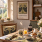 A styled dining room scene with a wooden table set for breakfast, featuring matching table linens, mugs, bowls, and a chair cushion with a black swan design on a beige and olive pattern. A framed William Morris-style print hangs on the wall above, and a window with warm sunlight is visible on the left.
