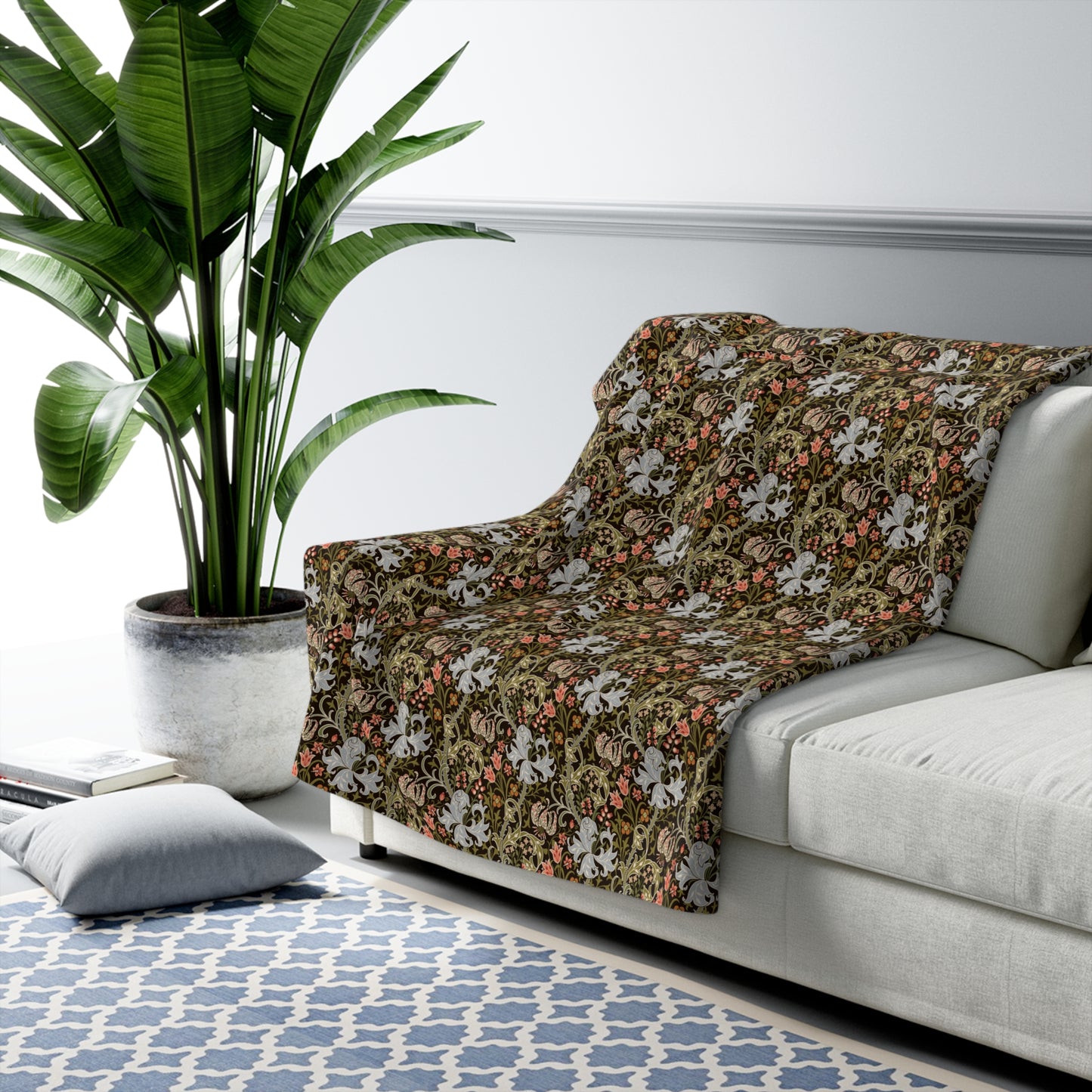 A floral throw blanket with a vintage botanical pattern draped over a light gray sofa in a modern living room setting next to a large potted plant.