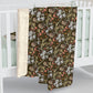 A baby blanket draped over a white crib, showing a dark brown and olive green floral and botanical pattern with white flowers and coral accents, and a soft cream sherpa backing.
