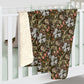 A folded baby blanket draped over a white crib, showing a dark brown and olive green floral pattern with white lilies and pink accents, alongside a soft cream sherpa fleece backing.