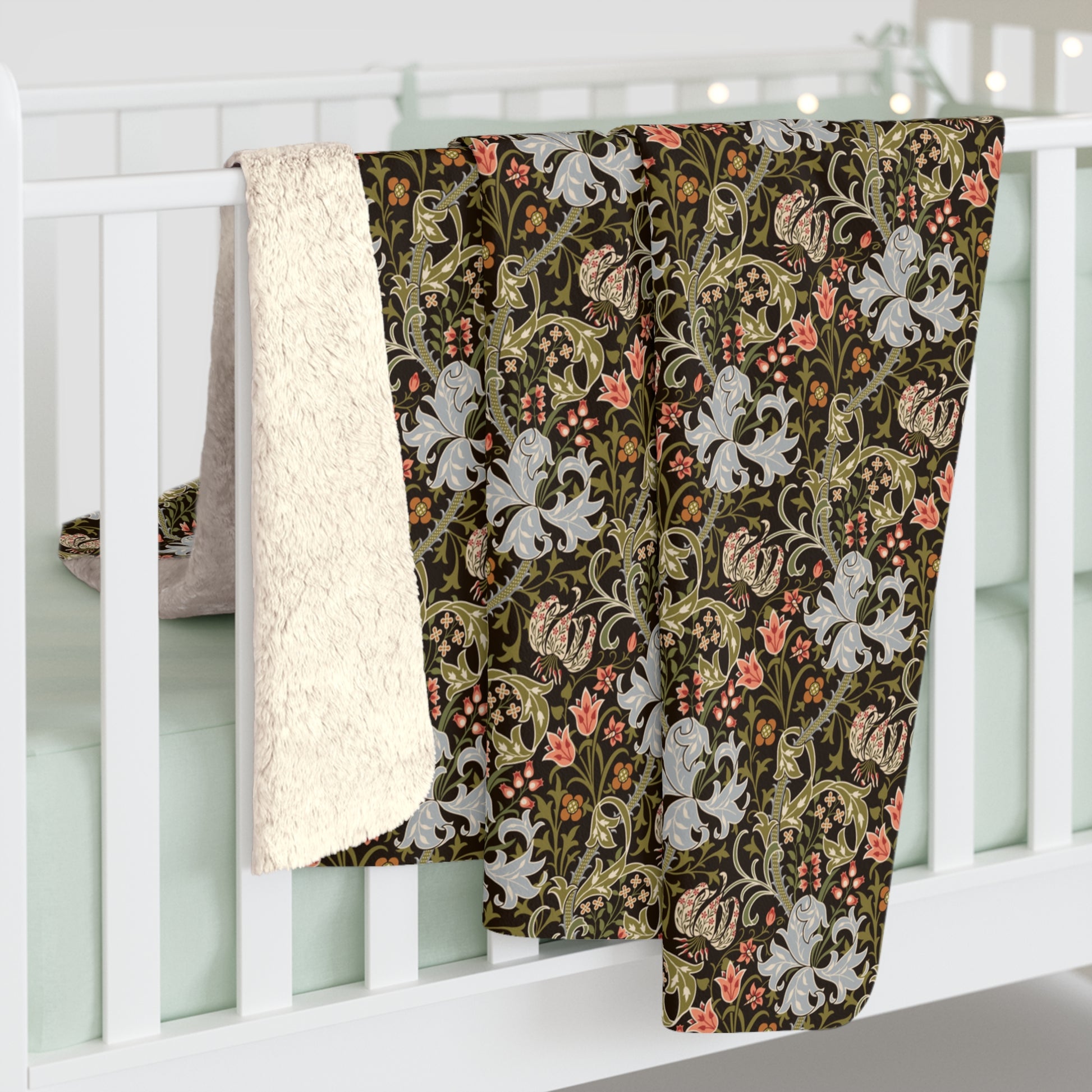 A folded baby blanket draped over a white crib, showing a dark brown and olive green floral pattern with white lilies and pink accents, alongside a soft cream sherpa fleece backing.