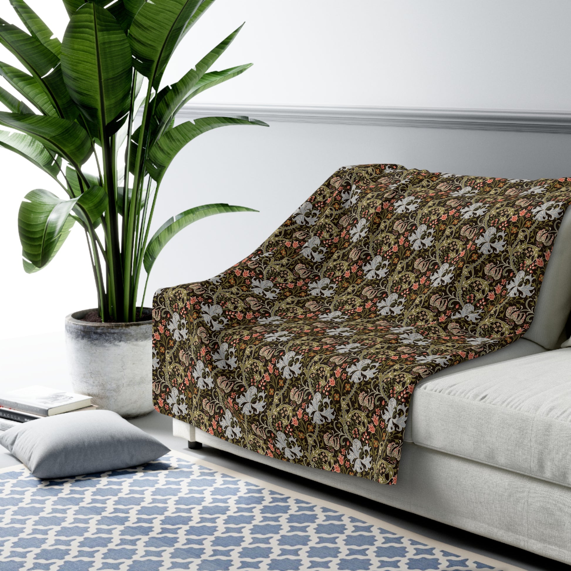 A floral throw blanket draped over a light gray sofa in a styled living room setting, featuring a dense botanical pattern in olive green, rust orange, and cream on a dark background.