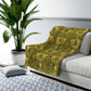 Green patterned throw blanket on a gray sofa with a plant and rug in the background