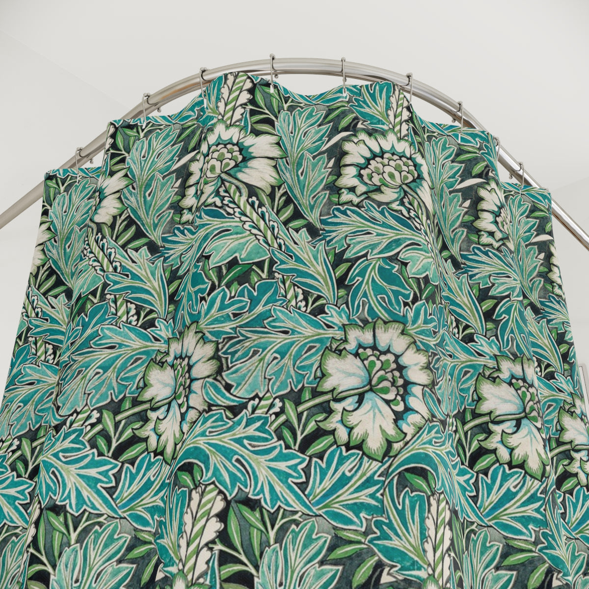 A bathroom scene showing a shower curtain with a teal, green, and white botanical leaf pattern hanging in a shower. The bathroom also features a white vanity with a marble countertop, a mirror, and a blue-gray accent wall.