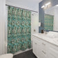 A bathroom scene showing a shower curtain with a teal, green, and white botanical leaf pattern hanging in a shower. The bathroom also features a white vanity with a marble countertop, a mirror, and a blue-gray accent wall.