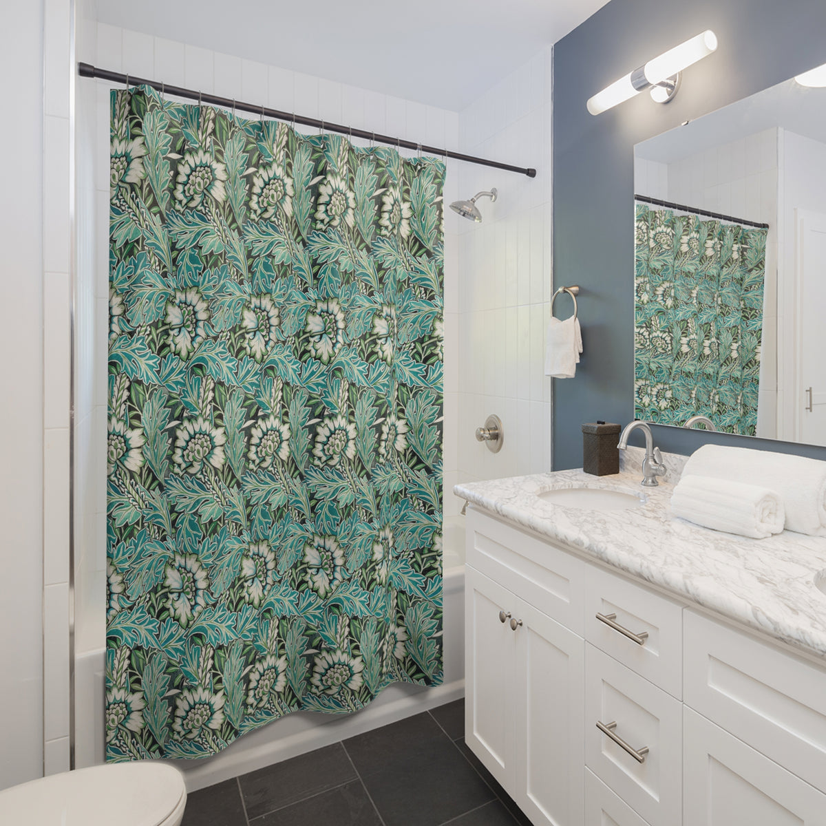 A bathroom scene showing a shower curtain with a teal, green, and white botanical leaf pattern hanging in a shower. The bathroom also features a white vanity with a marble countertop, a mirror, and a blue-gray accent wall.