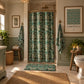 A bathroom scene showing a shower curtain with a teal, green, and white botanical leaf pattern hanging in a shower. The bathroom also features a white vanity with a marble countertop, a mirror, and a blue-gray accent wall.