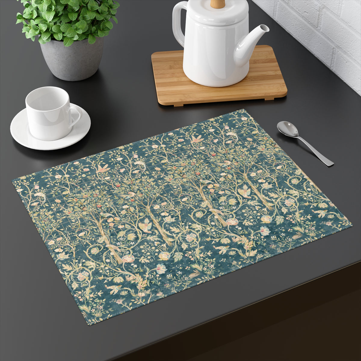 A rectangular table placemat with a vintage-style floral and bird pattern in teal and beige, styled on a dark tabletop with a white teapot, teacup, spoon, and potted plant.