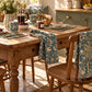 A rustic wooden dining table set in a sunlit kitchen, styled with matching blue floral tablecloths draped over the table and hanging from the chairs, along with ceramic mugs, a coffee pot, and breakfast dishes.