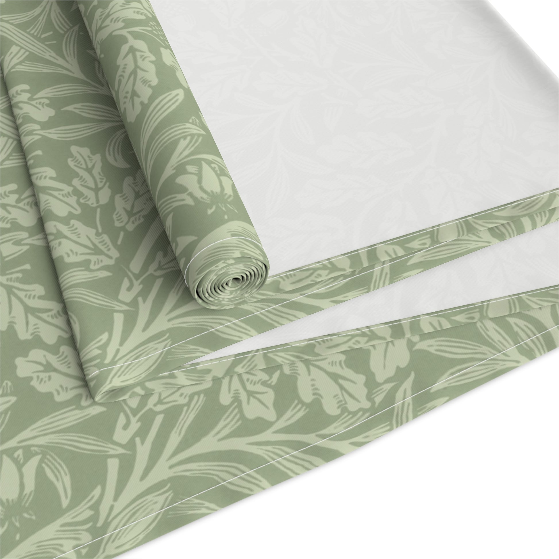 A green table runner with a floral William Morris & Co pattern, featuring acorns and oak leaves, laid out on a wooden dining table with cutlery and a vase of pink flowers in the background. 