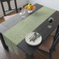 A green table runner with a floral William Morris & Co pattern, featuring acorns and oak leaves, laid out on a wooden dining table with cutlery and a vase of pink flowers in the background. 