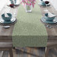 A green table runner with a floral William Morris & Co pattern, featuring acorns and oak leaves, laid out on a wooden dining table with cutlery and a vase of pink flowers in the background. 