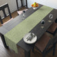 A green table runner with a floral William Morris & Co pattern, featuring acorns and oak leaves, laid out on a wooden dining table with cutlery and a vase of pink flowers in the background. 