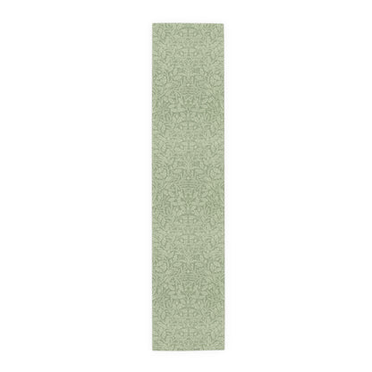A green table runner with a floral pattern, featuring acorns and oak leaves, laid out on a wooden dining table with cutlery and a vase of pink flowers in the background.
