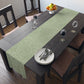 A green table runner with a floral William Morris & Co pattern, featuring acorns and oak leaves, laid out on a wooden dining table with cutlery and a vase of pink flowers in the background. 