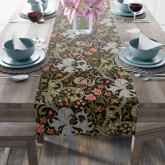 A table runner with a floral William Morris & Co pattern in shades of green, gold, and other colours, laid out on a dining table with dishes and glasses arranged on top.
