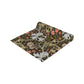 A table runner with a floral William Morris & Co pattern in shades of green, gold, and other colours, laid out on a dining table with dishes and glasses arranged on top.