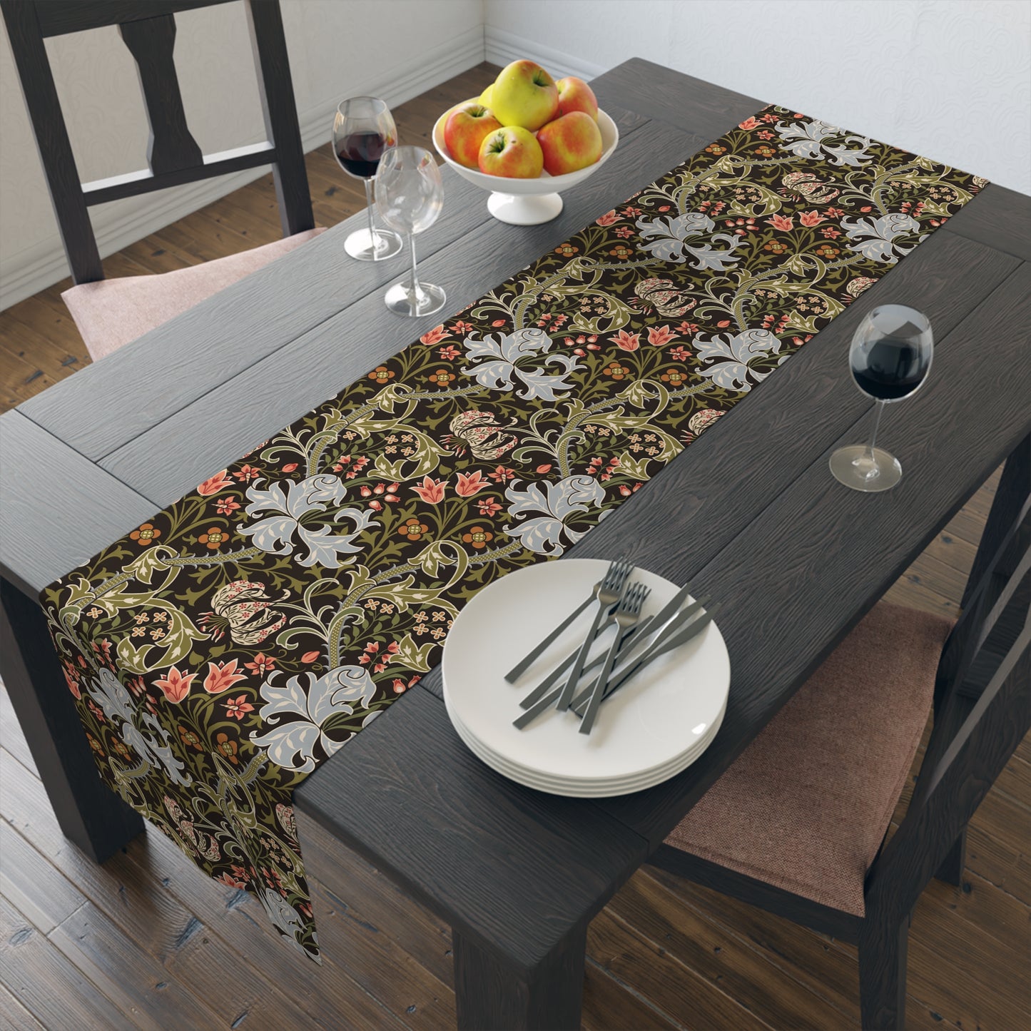 A table runner with a floral William Morris & Co pattern in shades of green, gold, and other colours, laid out on a dining table with dishes and glasses arranged on top.