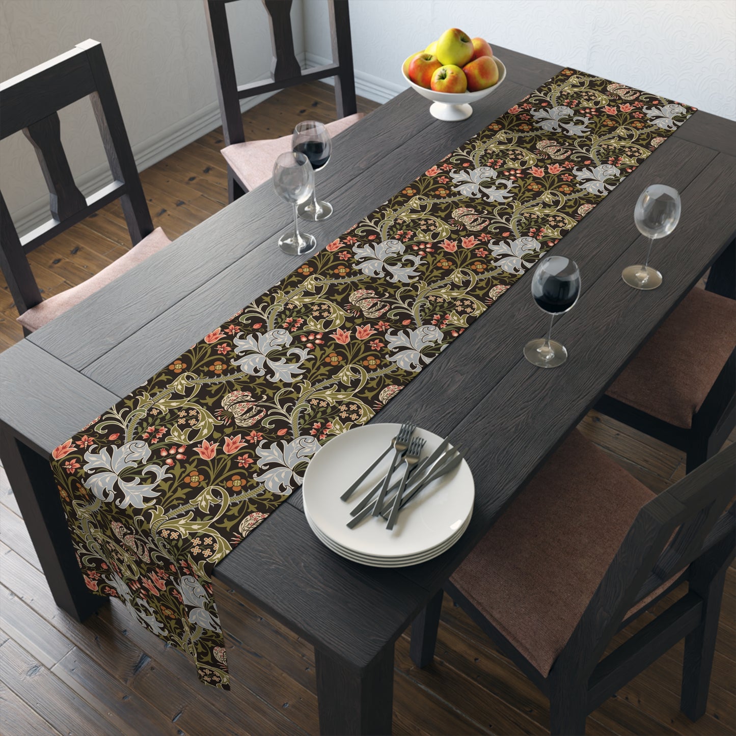 A table runner with a floral William Morris & Co pattern in shades of green, gold, and other colours, laid out on a dining table with dishes and glasses arranged on top.