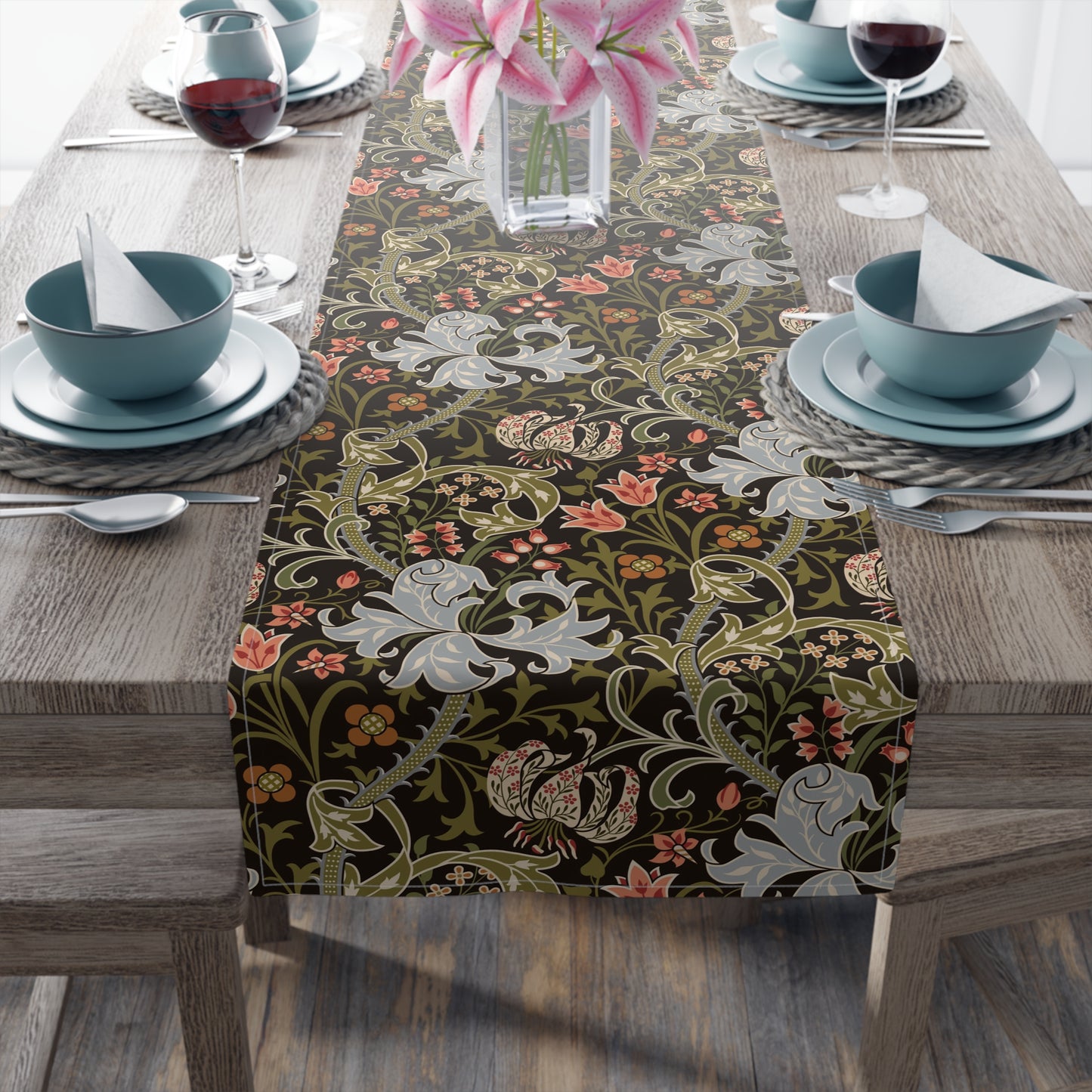 A table runner with a floral pattern in shades of green, gold, and other colors, laid out on a dining table with dishes and glasses arranged on top.