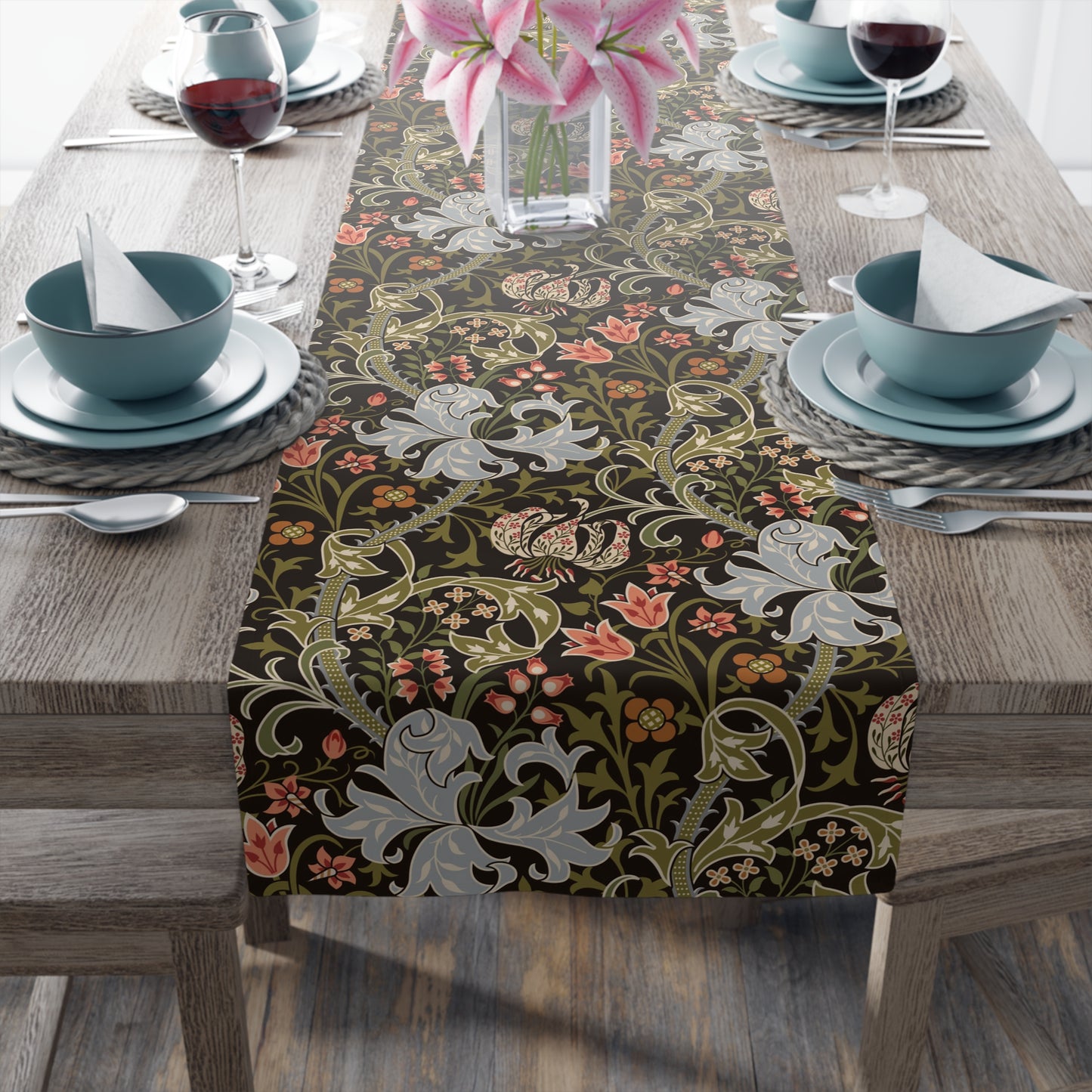 A table runner with a floral pattern in shades of green, gold, and other colors, laid out on a dining table with dishes and glasses arranged on top.