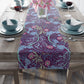 A multicolored table runner with a floral William Morris & Co pattern in pink and green on a blue background, displayed on a table with dishes and cutlery.