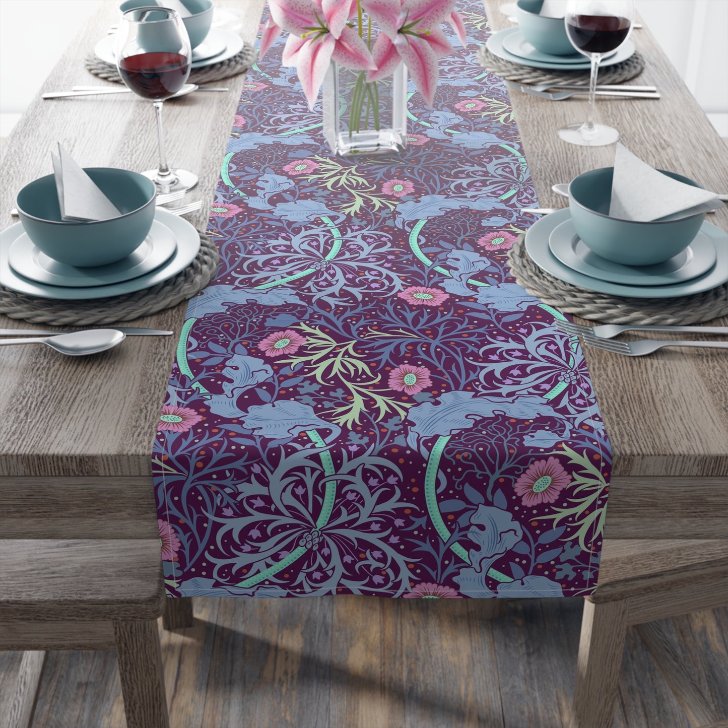 A multicolored table runner with a floral pattern in pink and green on a blue background, displayed on a table with dishes and cutlery.