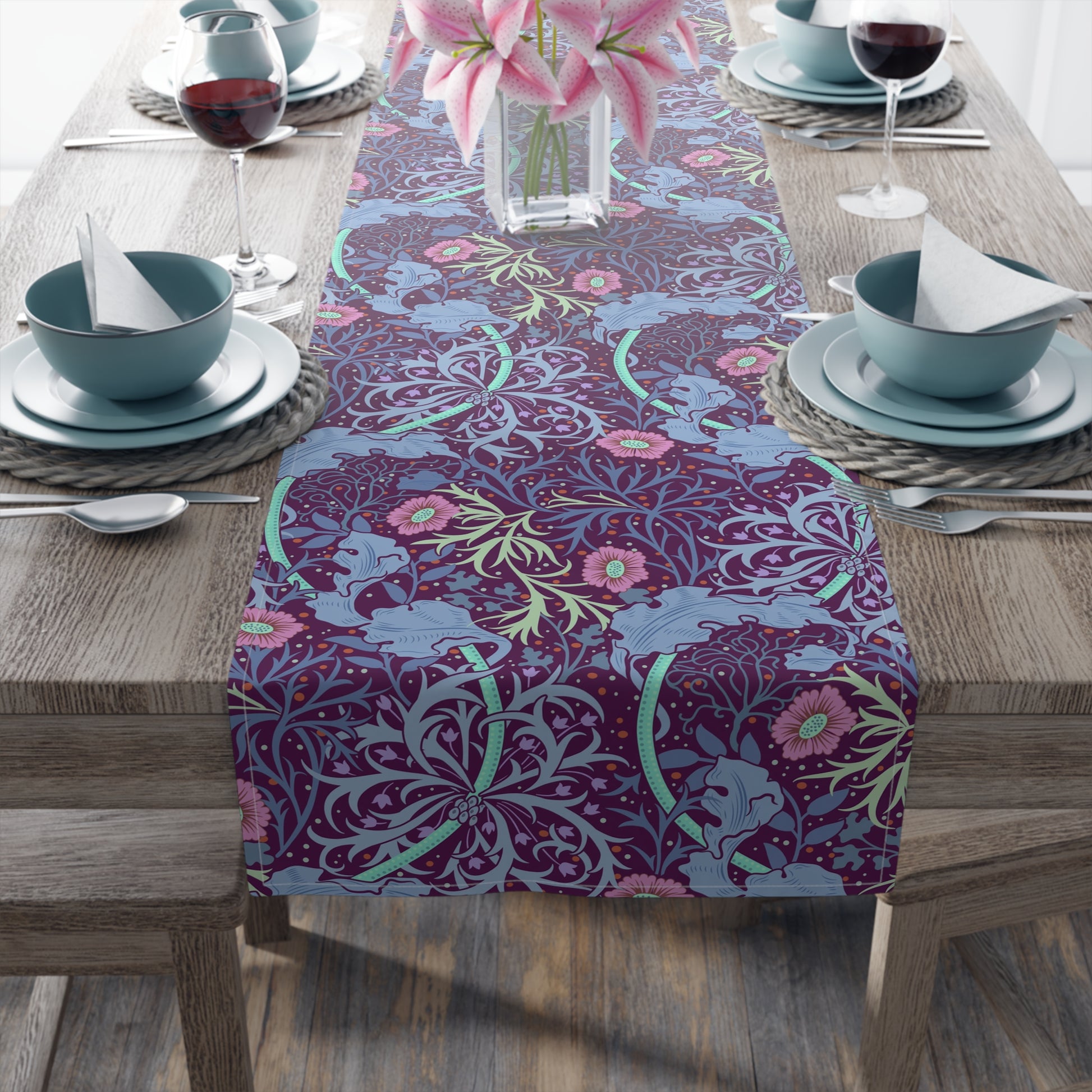 A multicolored table runner with a floral pattern in pink and green on a blue background, displayed on a table with dishes and cutlery.