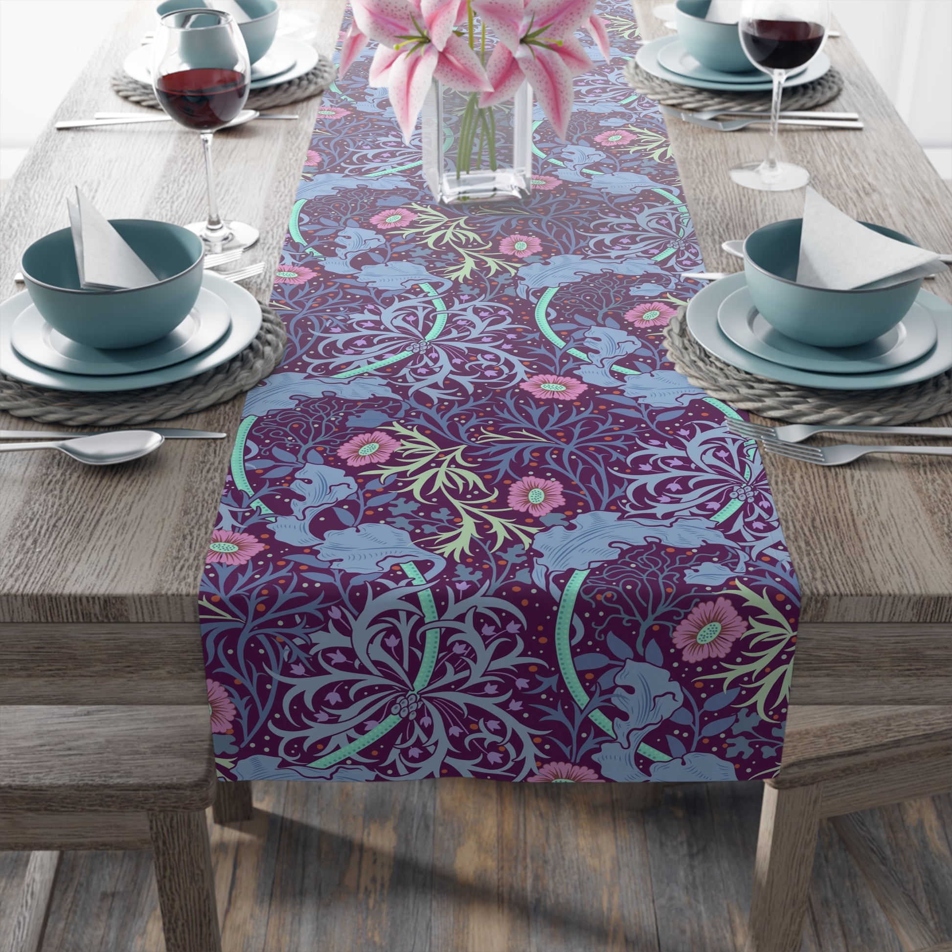 A multicolored table runner with a floral pattern in pink and green on a blue background, displayed on a table with dishes and cutlery.