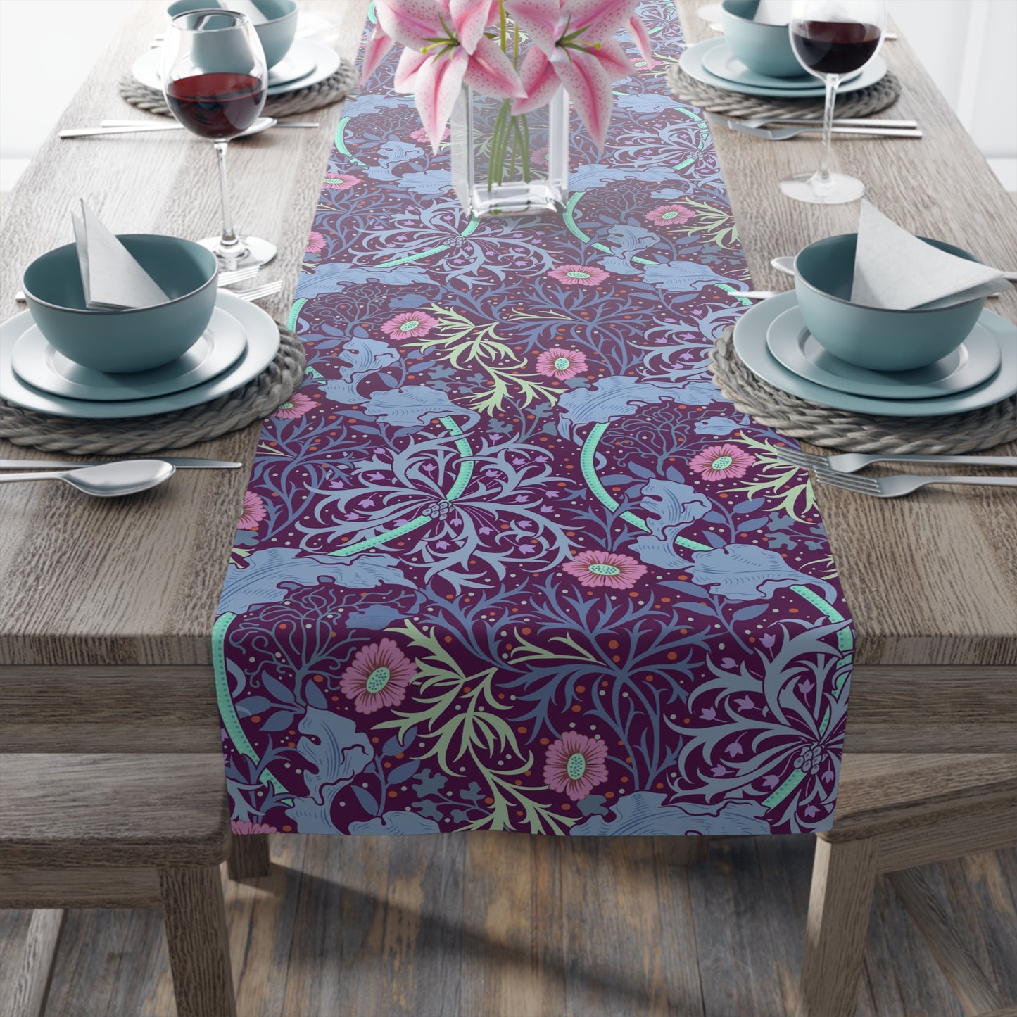A multicolored table runner with a floral pattern in pink and green on a blue background, displayed on a table with dishes and cutlery.